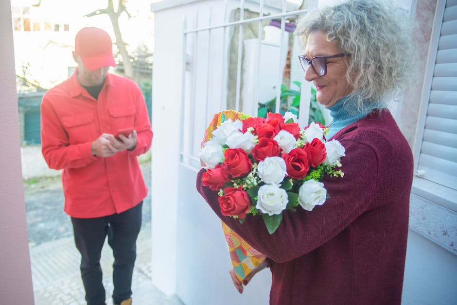 A woman with curly gray hair wearing glasses and a burgundy sweater holding a bouquet of fresh red and white roses with green foliage, wrapped in colorful paper, during a flower delivery at her home. 