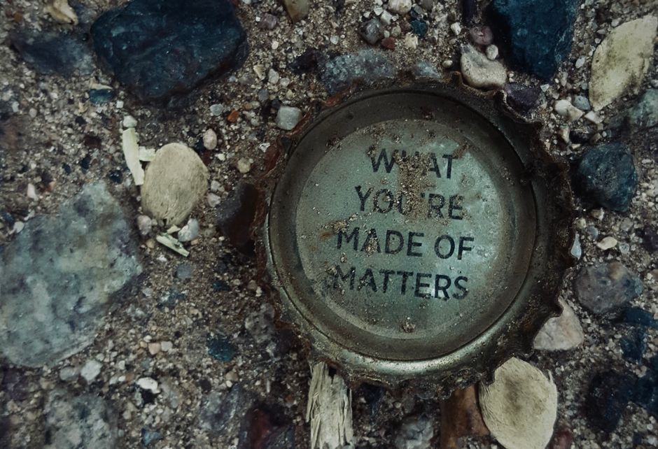 A close-up of a round metal bottle cap lying on a gravel surface, with the phrase 'WHAT YOU'RE MADE OF MATTERS' printed in black capital letters on its top. The cap appears weathered and slightly rust