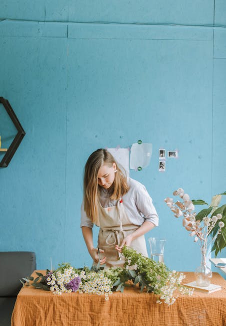 A young woman with shoulder-length blonde hair, wearing a light gray shirt and beige apron, stands at a table arranging an assortment of fresh flowers, including white daisies, pale pink and purple bl