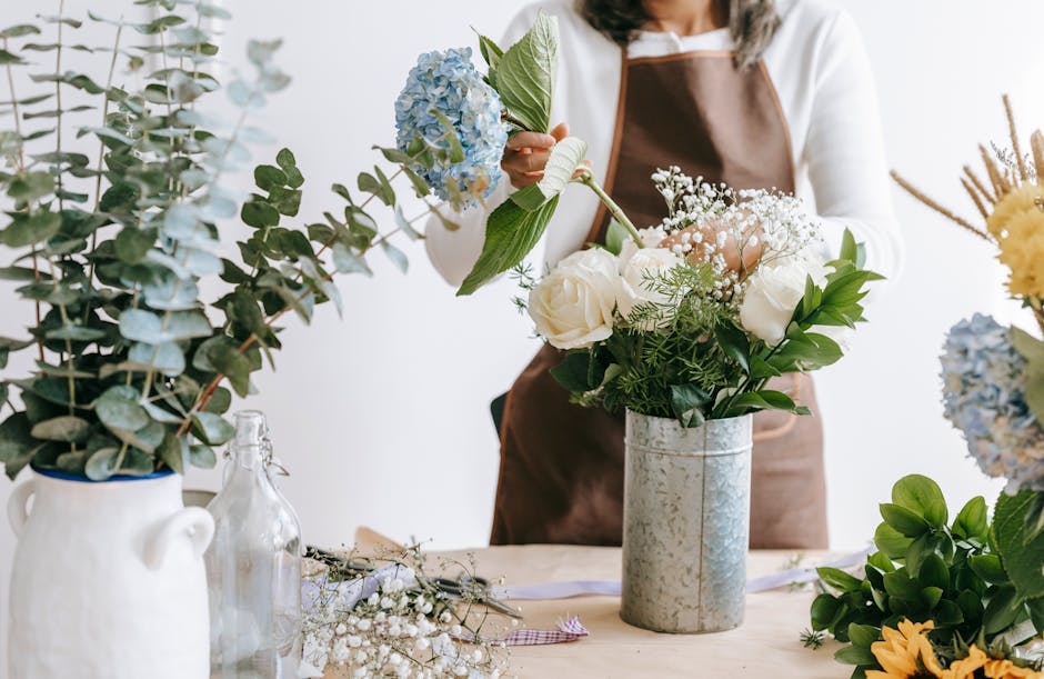 A florist wearing a brown apron and a white long-sleeve shirt is arranging a floral bouquet in a tall, cylindrical, textured silver vase on a wooden table. The bouquet features large white roses, spri