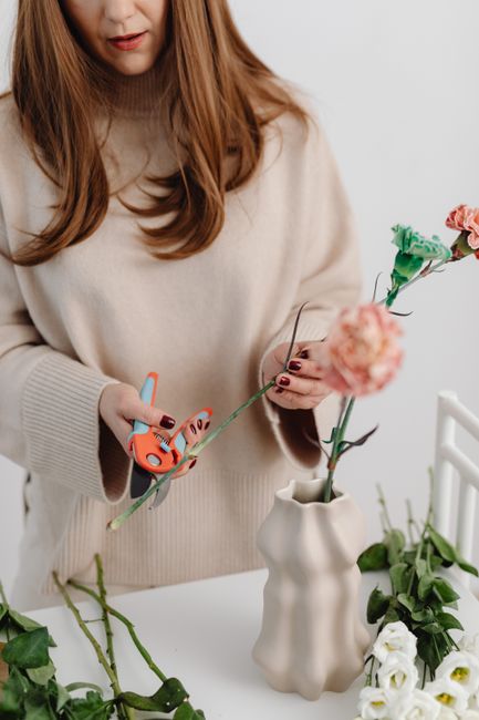 A woman with long auburn hair wearing a beige sweater is holding a pair of small, orange-handled scissors in her right hand and a length of green floral wire in her left. She is assembling a floral ar