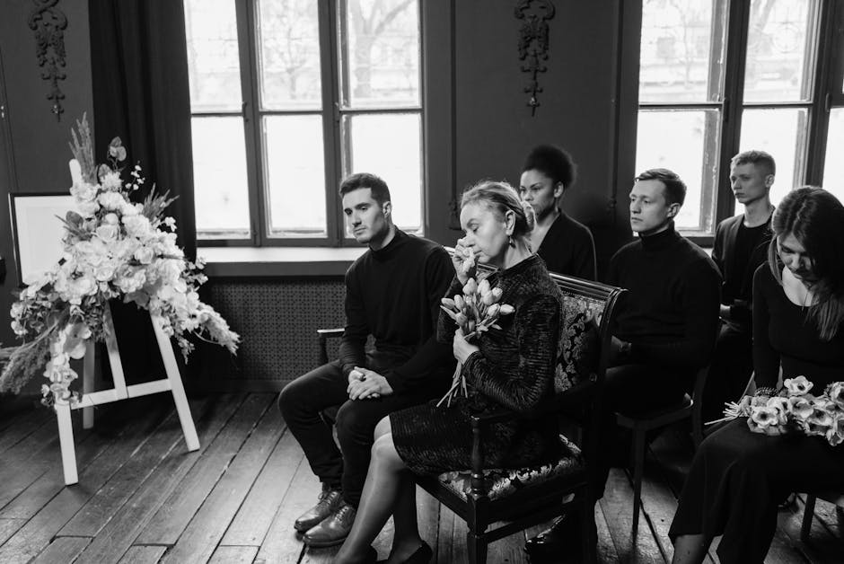 A black and white photograph showing a group of six people seated inside a somber room with high windows, during a funeral service. The central figure, an elderly woman with short hair, holds a bouque