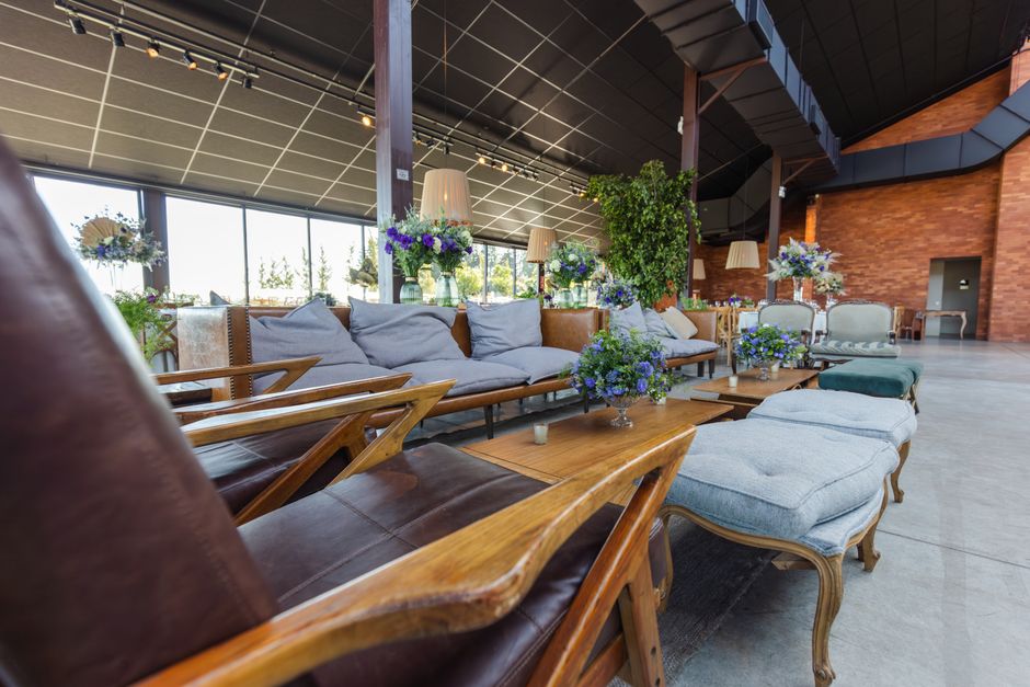 A spacious, modern lounge area with large floor-to-ceiling windows allowing natural light to illuminate the space. The room features a variety of floral arrangements, including bouquets of blue and pu