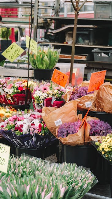 A vibrant outdoor flower stall displays various floral arrangements and bouquets wrapped in brown paper, showcasing a mix of fresh flowers including pink tulips, white daisies, purple lavender, and gr