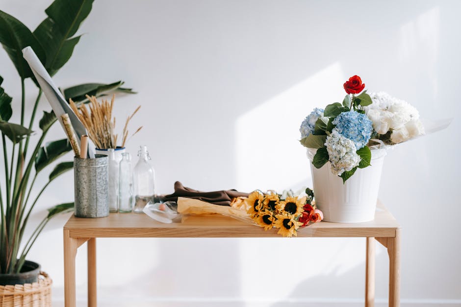 A white ceramic vase on a light wooden table holding a floral arrangement of white hydrangeas, blue hydrangeas, and a single red rose with green foliage, set in a well-lit space with natural sunlight.