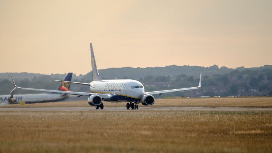 A Ryanair commercial airplane with a white fuselage and blue and yellow livery on the tail, taxiing on a runway with a grassy foreground. The sky is overcast with a soft, cloudy appearance, and in the