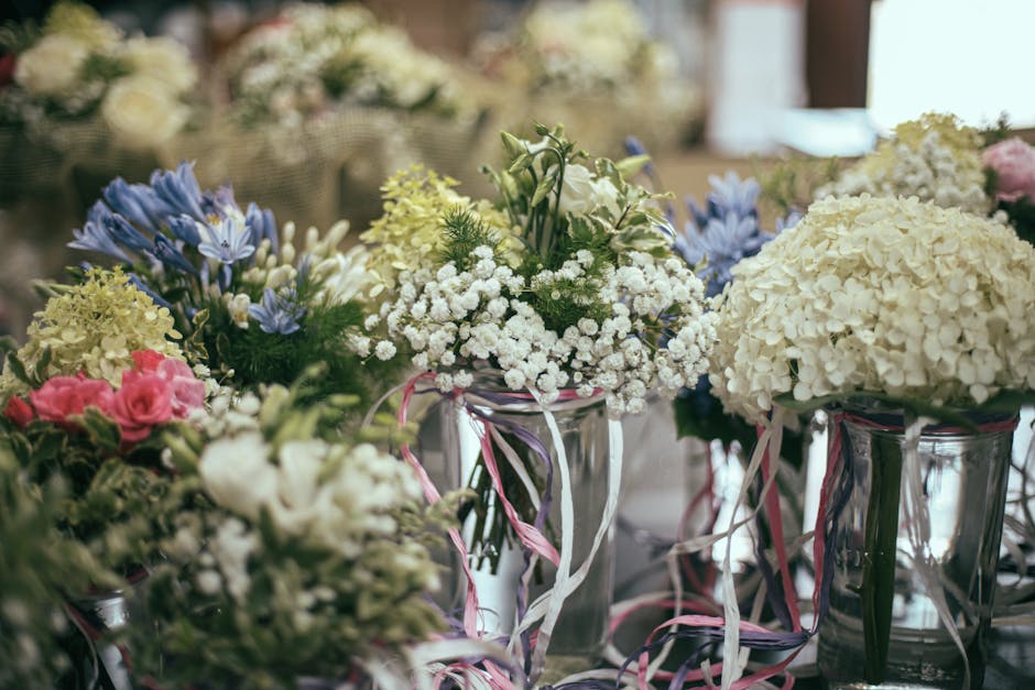 A collection of floral arrangements featuring white hydrangeas, pink roses, bluebells, and sprigs of green foliage, displayed in clear glass vases decorated with pink and white ribbon streamers. The f